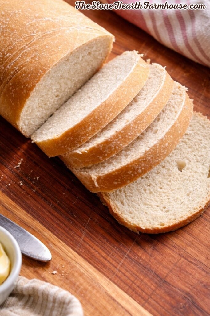 Freshly baked sourdough sandwich bread sliced on a wooden cutting board with a small dish of butter, bread knife, and farmhouse-style kitchen towel in the background.
