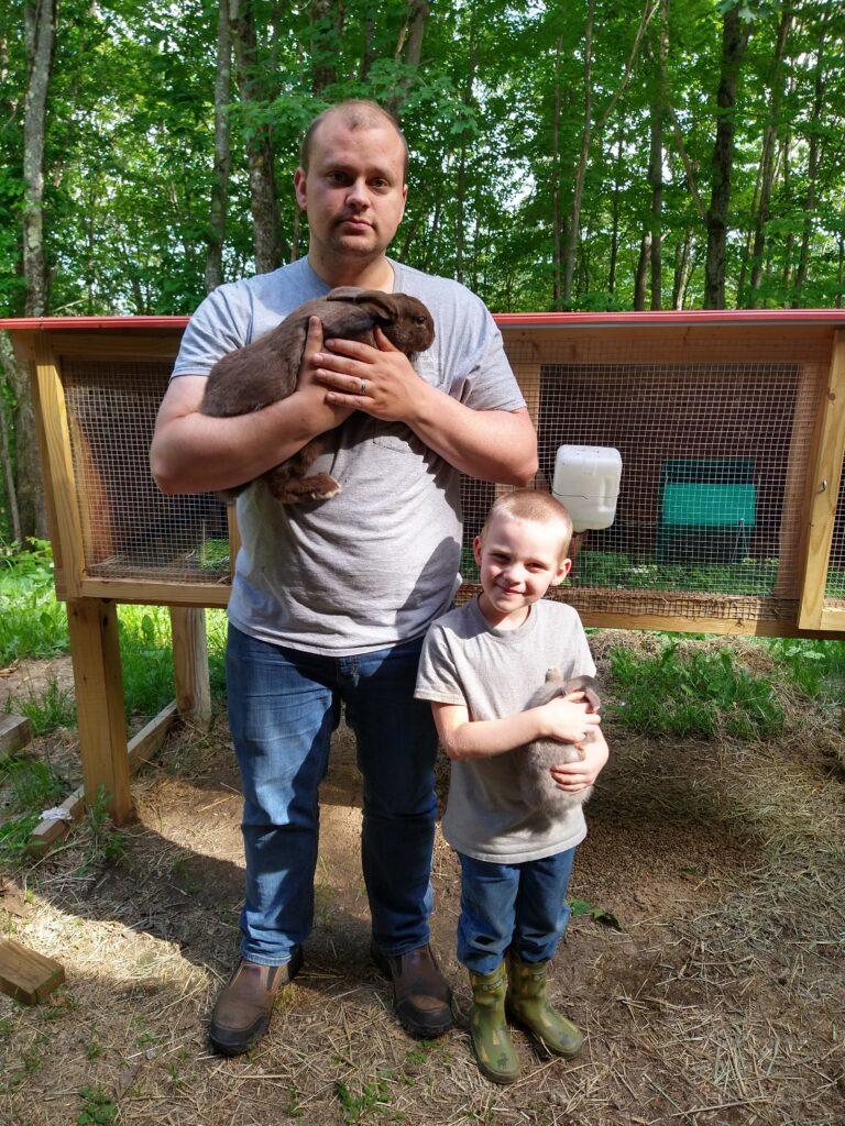 man and little boy standing in front of a rabbit hutch outside and both smiling and holding a rabbit