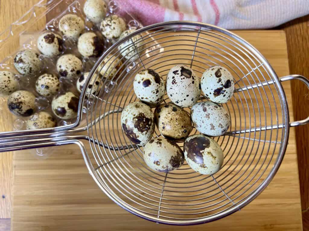 nine quail eggs in a slotted utensil with a carton of quail eggs in the back round. 