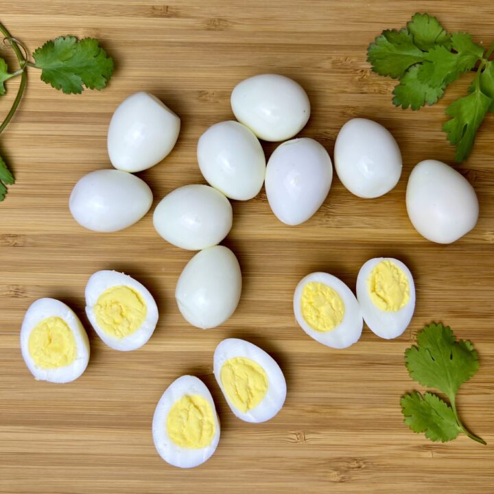 A group of hard-boiled quail eggs, some whole and some halved to show the yolks, arranged on a wooden cutting board with fresh cilantro leaves for garnish.