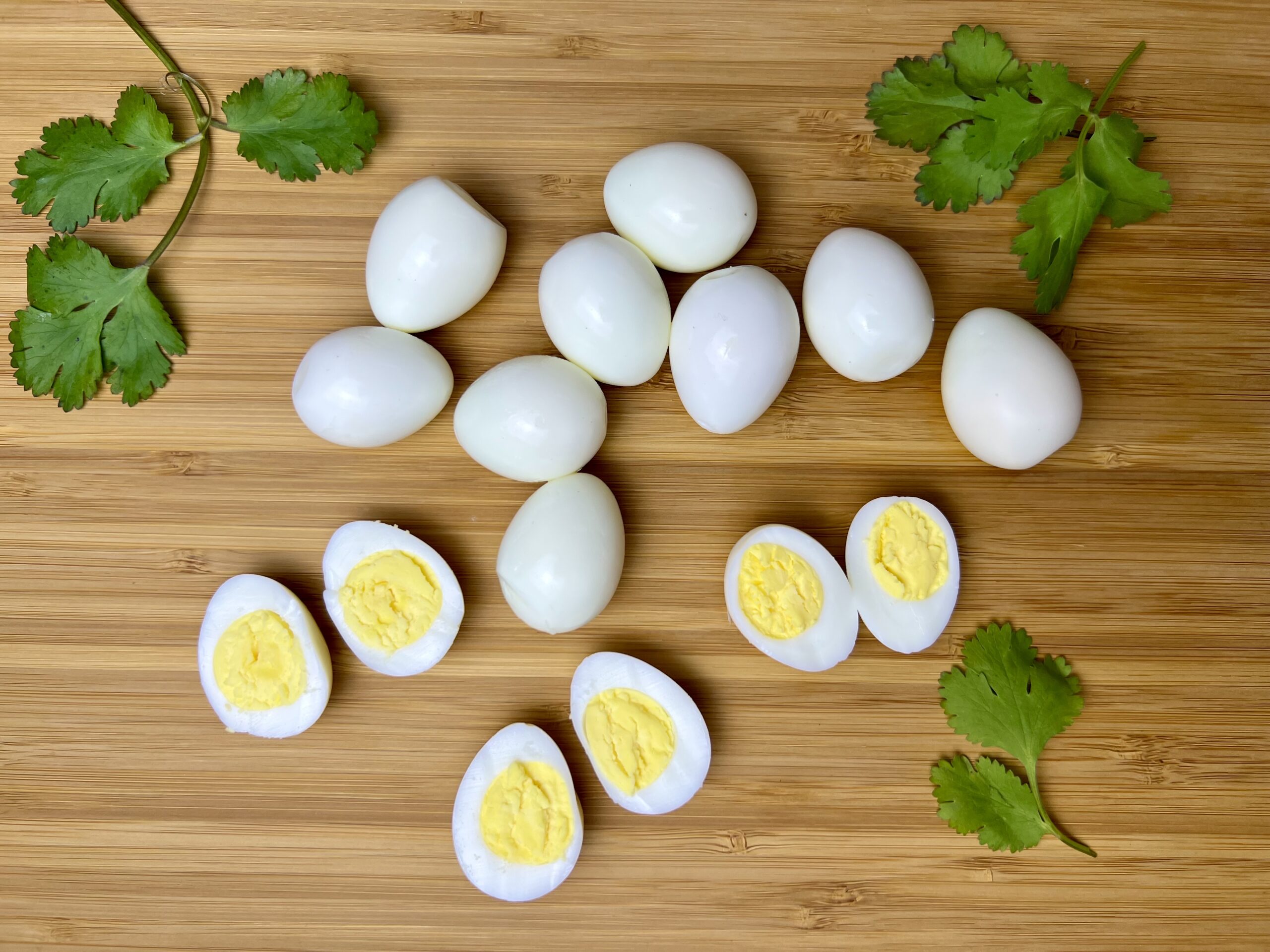A group of hard-boiled quail eggs, some whole and some halved to show the yolks, arranged on a wooden cutting board with fresh cilantro leaves for garnish.