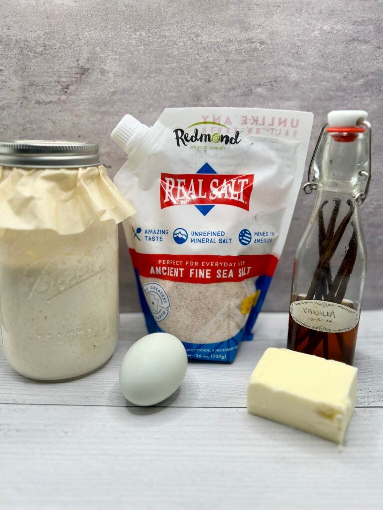 Ingredients for sourdough discard pancakes including sourdough starter, egg, butter, vanilla extract, and sea salt on a rustic kitchen surface.