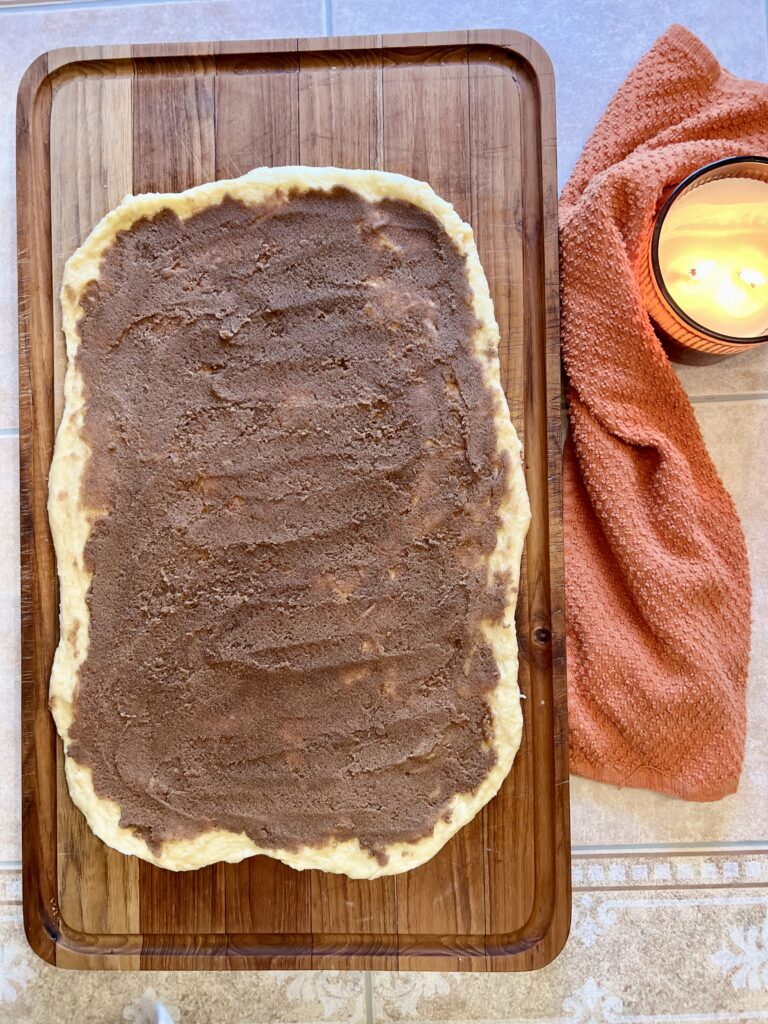 Sourdough cinnamon roll dough rolled out and spread evenly with brown sugar cinnamon filling on a wooden board.