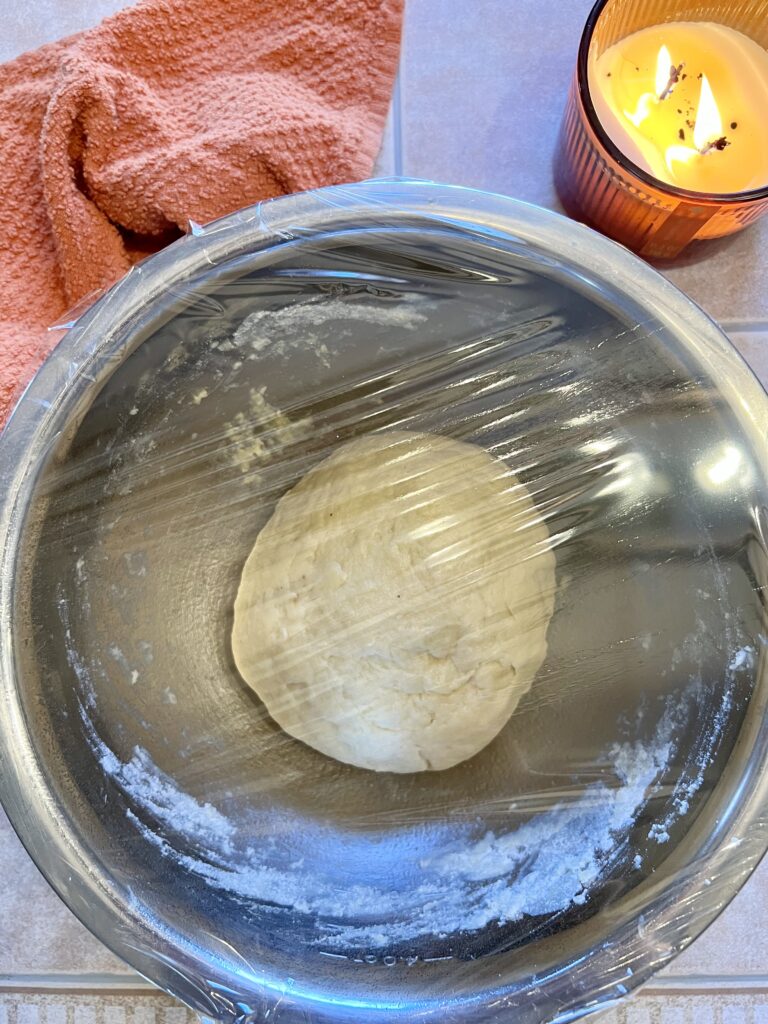Sourdough cinnamon roll dough rising in a glass bowl covered with plastic wrap during overnight fermentation.