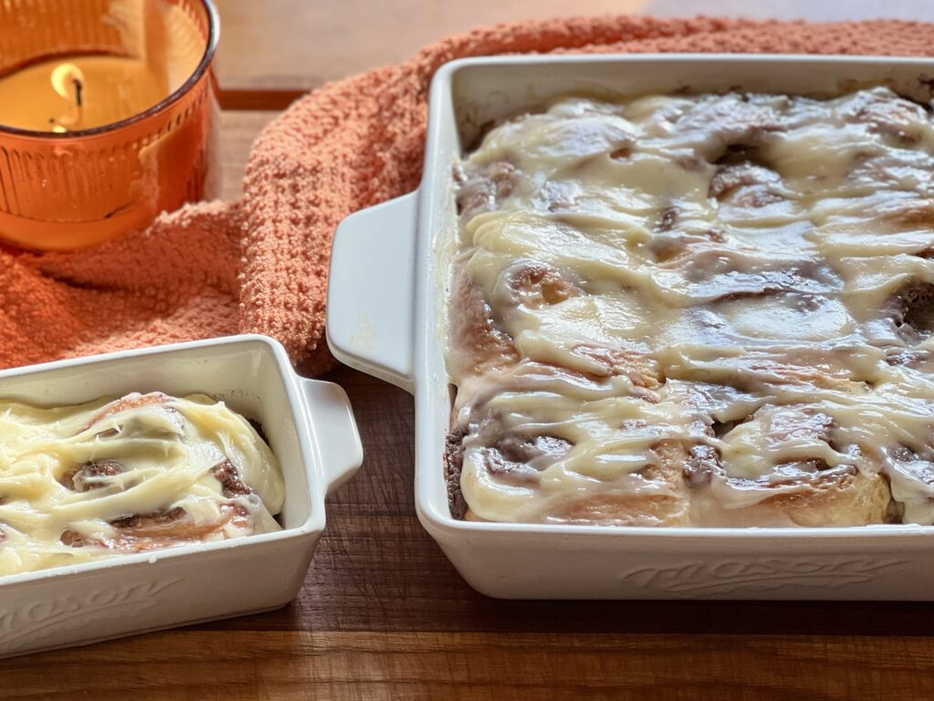 Freshly baked sourdough cinnamon rolls in a white baking dish topped with thick cream cheese frosting, served on a wooden table with a cozy fall candle.