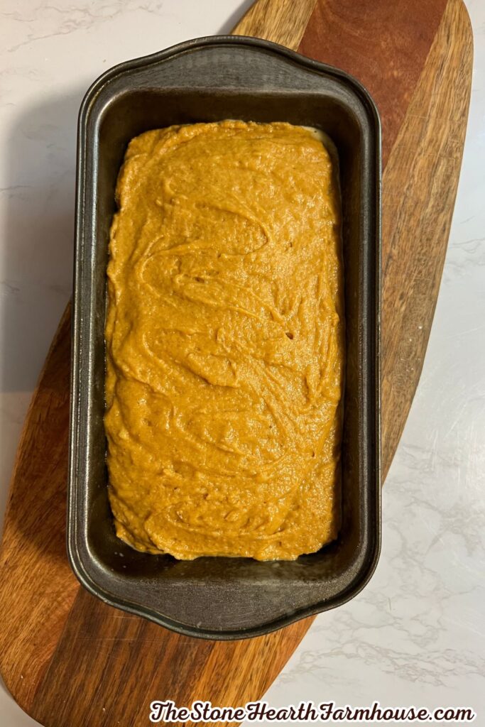 Pumpkin bread batter spread evenly in a dark loaf pan before baking, with a wooden cutting board underneath.