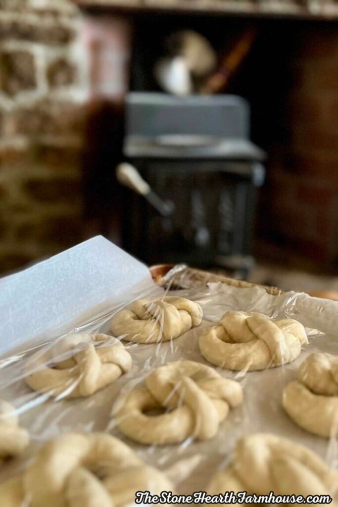Shaped pretzels resting under plastic wrap on a parchment-lined baking sheet