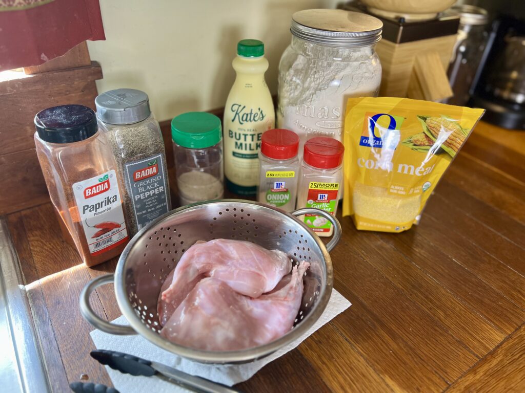 Ingredients for making fried rabbit including raw rabbit meat, cornmeal, flour, buttermilk, and seasonings like paprika, black pepper, garlic powder, and onion powder on a wooden kitchen counter