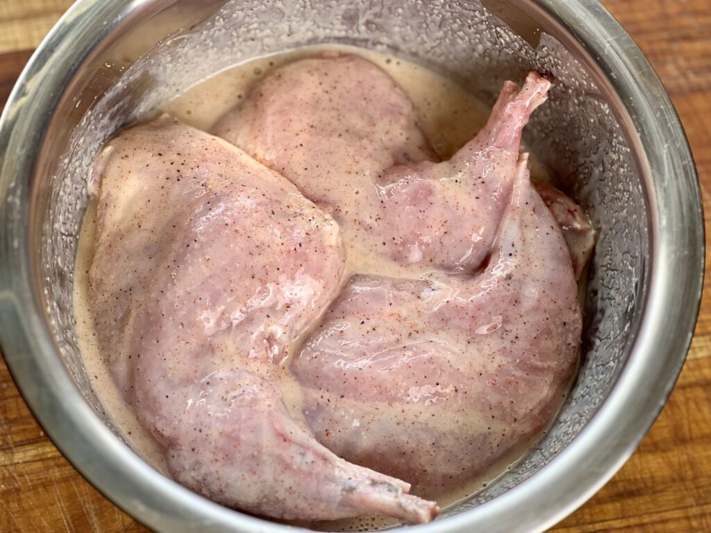 Raw rabbit meat marinating in seasoned buttermilk in a stainless steel bowl, preparing for Southern-style fried rabbit
