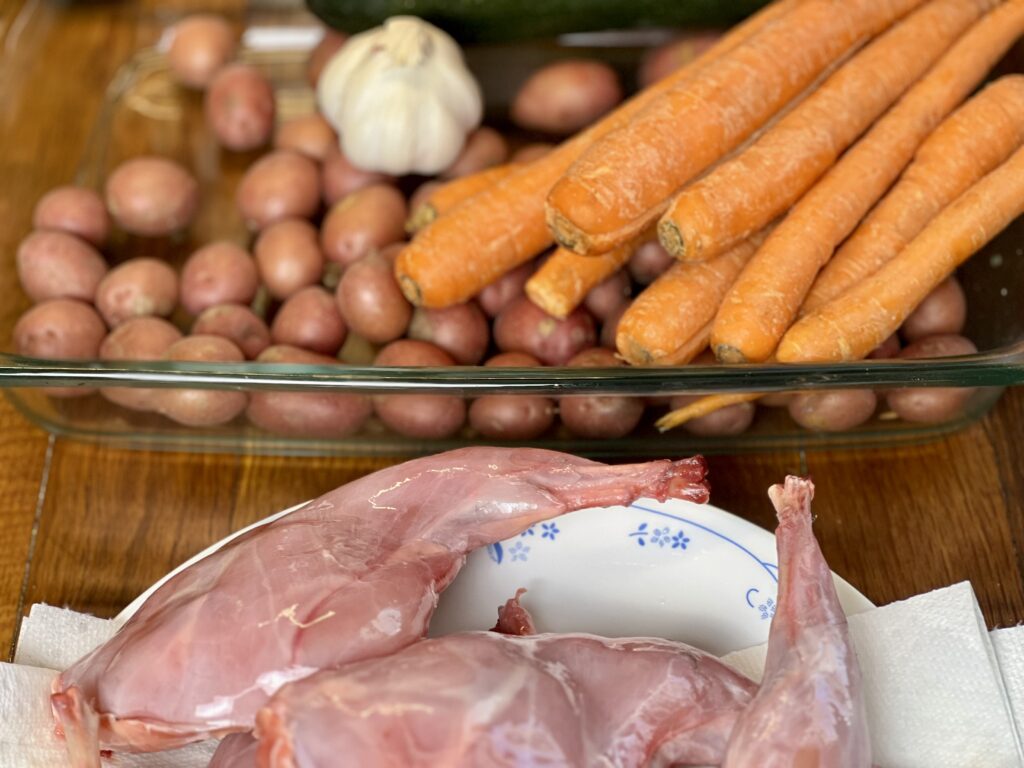 uncooked rabbit thighs on a plate in front of a pan of carrots and potatoes