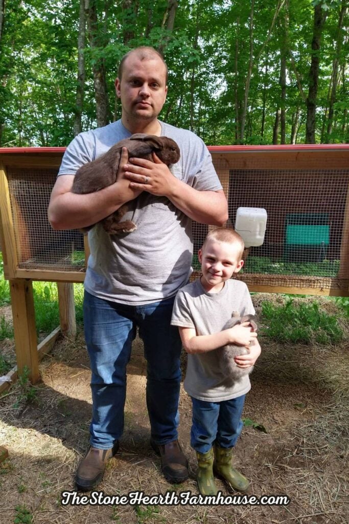 man and little boy standing in front of a rabbit hutch outside and both smiling and holding a rabbit