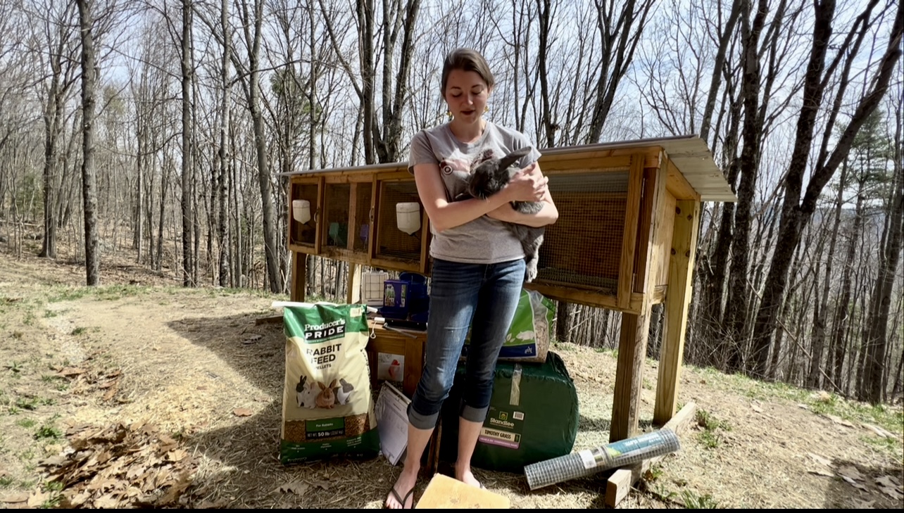 women holding a rabbit in front of a hutch with a display of everything a beginner needs to raise meat rabbits.
