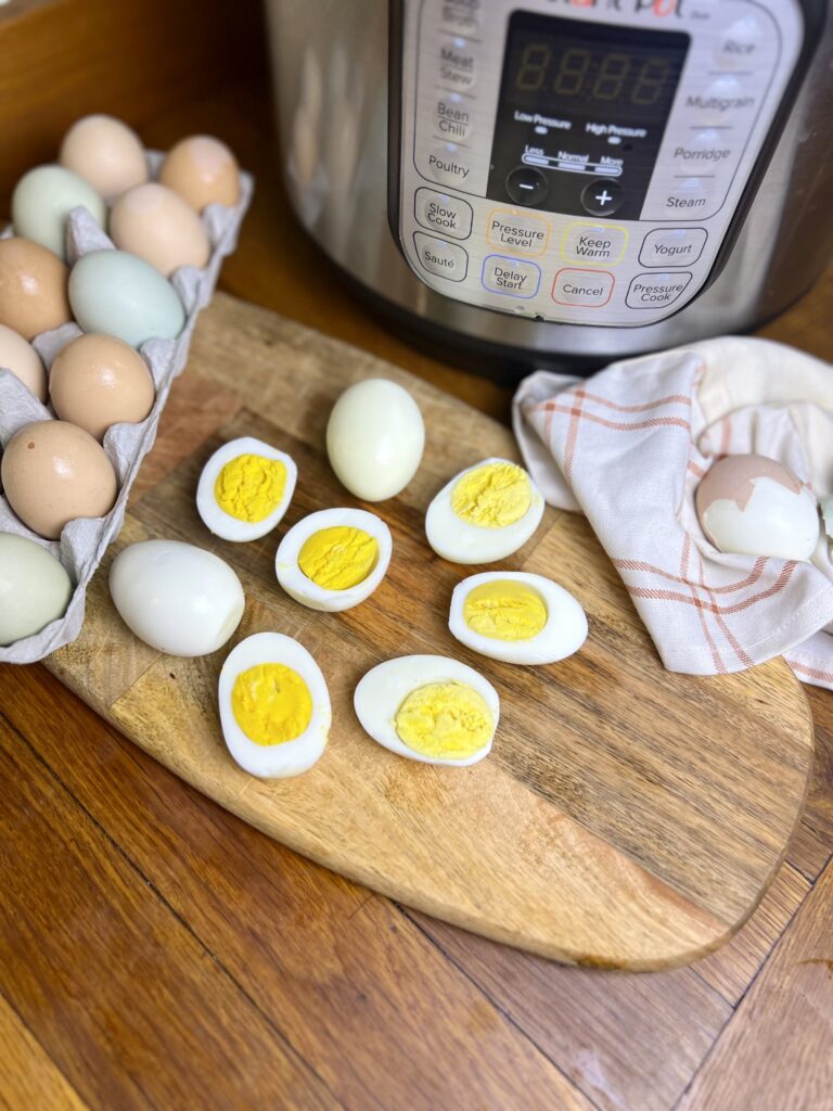 Hard boiled eggs cut in half on a wooden cutting board next to an Instant Pot, with a carton of farm-fresh brown and blue eggs in the background.
