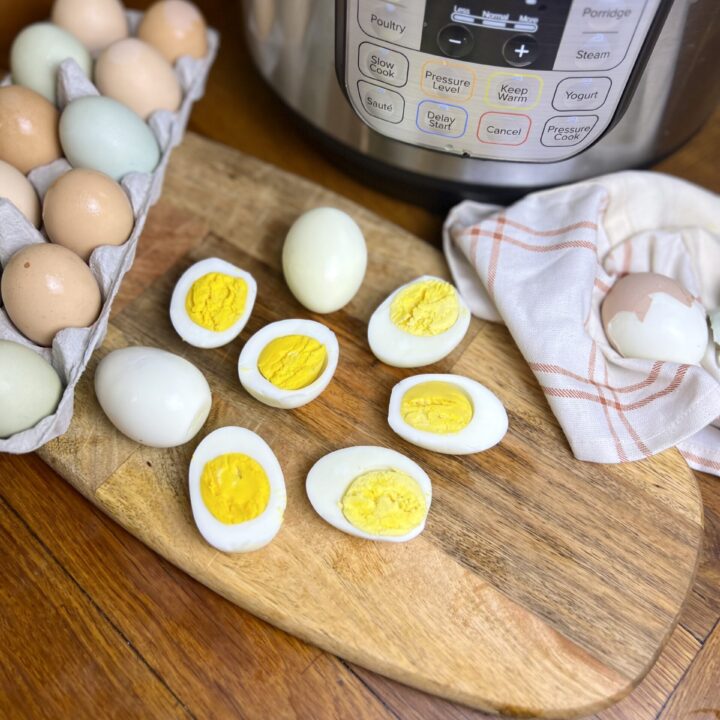Hard boiled eggs cut in half on a wooden cutting board next to an Instant Pot, with a carton of farm-fresh brown and blue eggs in the background.