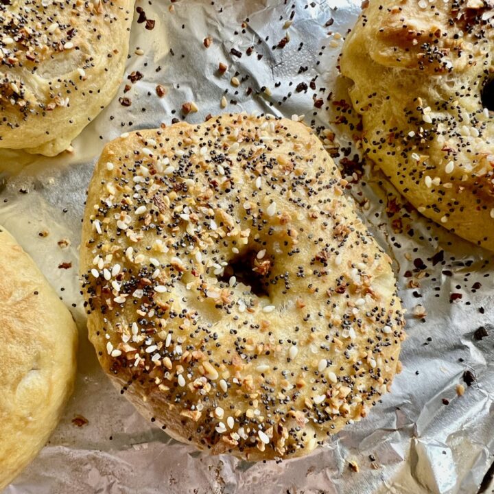 Freshly baked homemade sourdough bagels topped with everything bagel seasoning, resting on a foil-lined baking sheet with golden, chewy crusts and scattered seasoning crumbs.