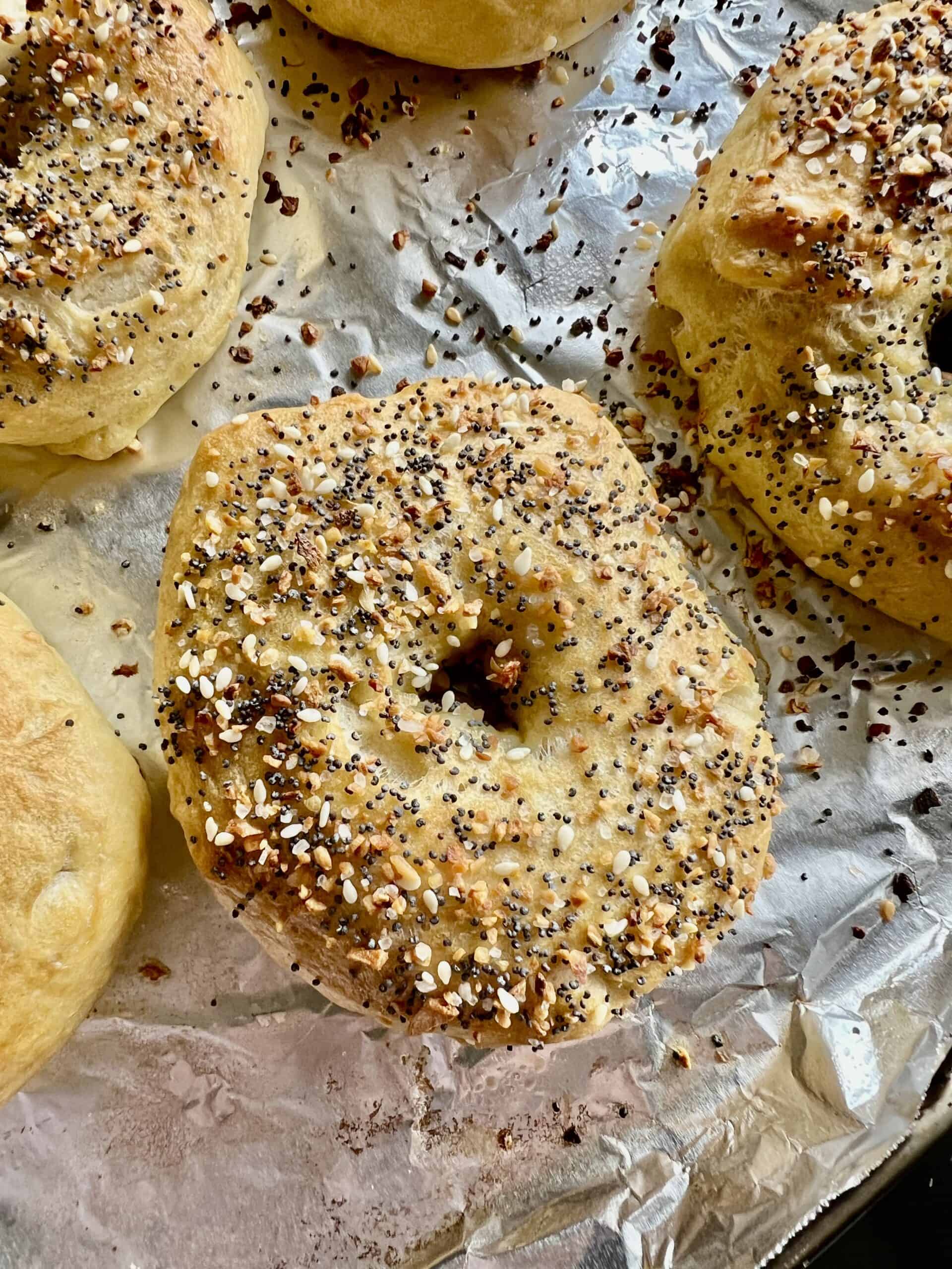 Freshly baked homemade sourdough bagels topped with everything bagel seasoning, resting on a foil-lined baking sheet with golden, chewy crusts and scattered seasoning crumbs.