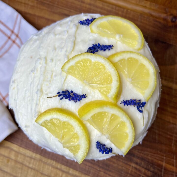 looking down at the top of a cake there is scliced lemmons and purple flowers decorating the buttercream frosting.