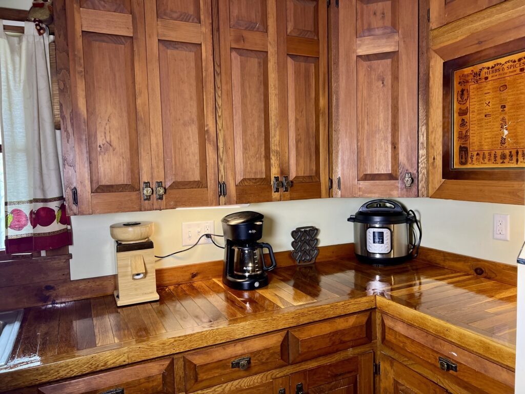 Shiny, handcrafted oak wood countertops made from hardwood flooring, paired with vintage-style wooden cabinets and modern appliances like a NutriMill grain mill, coffee maker, and pressure cooker.”
