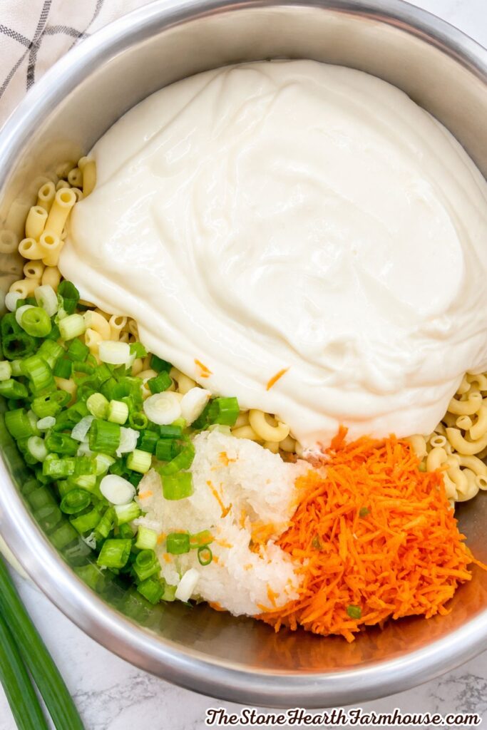 Cooked macaroni combined with creamy dressing, shredded carrots, grated onion, and green onions in a mixing bowl