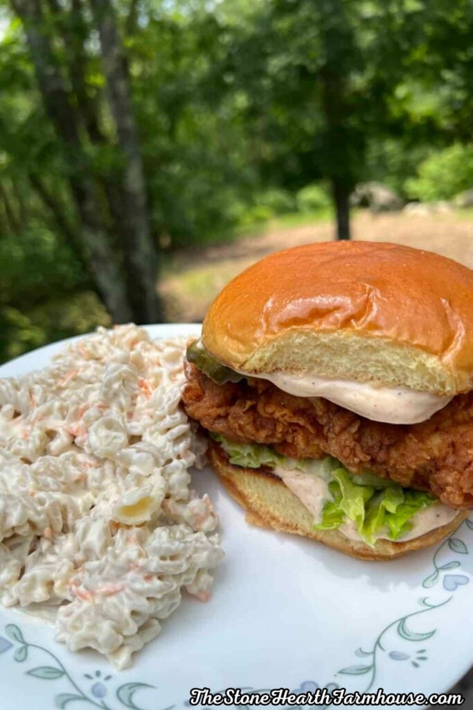 Crispy buttermilk chicken sandwich on a toasted brioche bun with lettuce, pickles, and creamy sauce, served with creamy pasta salad