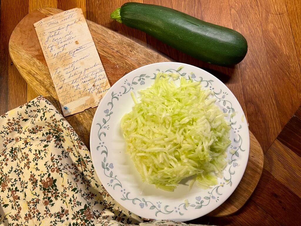 grated zucchini on a plate next to a old recipe card on a wooden cutting board.