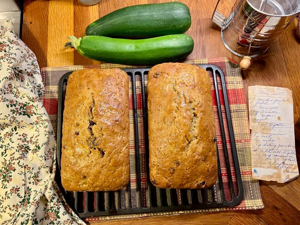 two fresh loaves of zucchini bread on a cast iron rack cooling next to two green zucchinis.