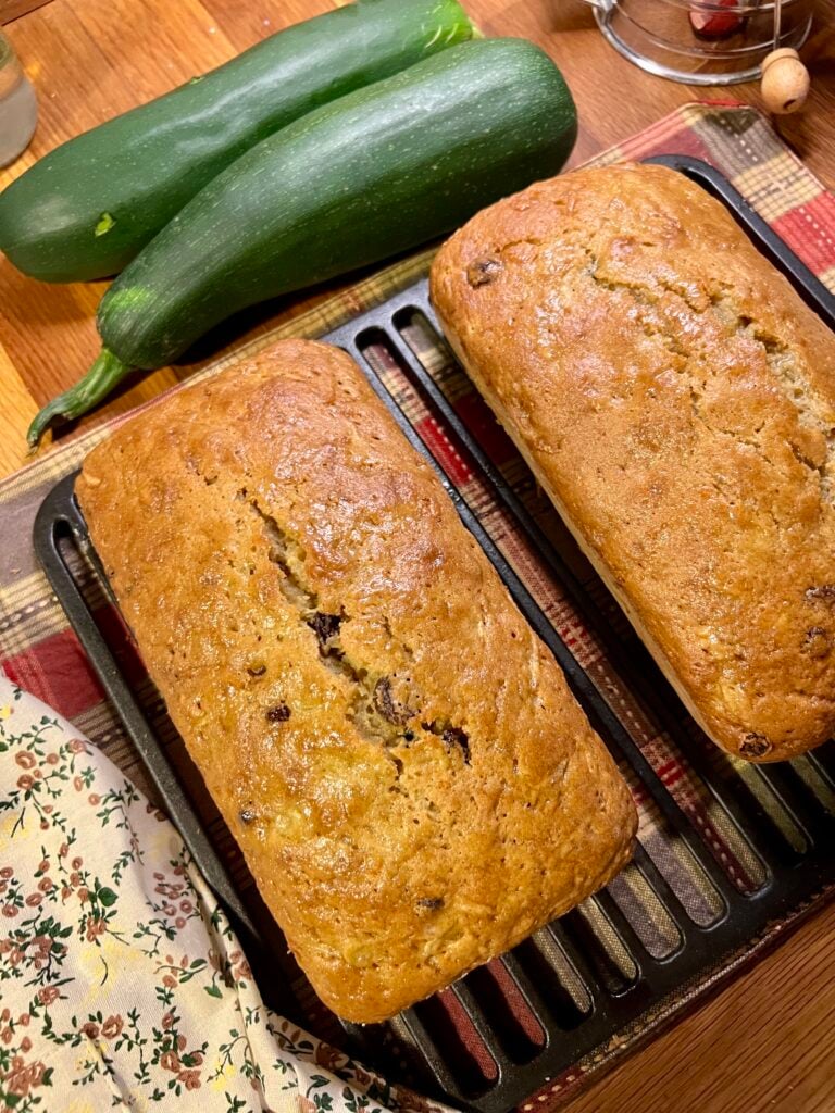 2 Loaves of freshly baked zucchini bread cooling next to two green zucchini.
