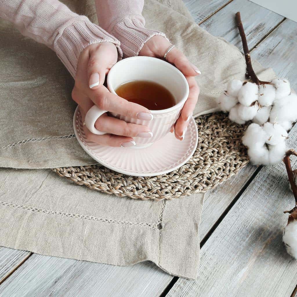Hands holding a warm cup of tea surrounded by rustic textiles and cotton.
