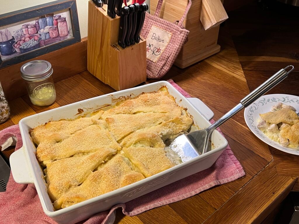 On a wooden counter tip sits the casserole dish with chicken crescent casserole on it. next to it is a piece of the casserole on a plate The casserole is being served.