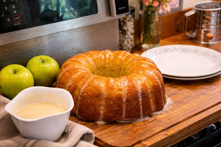 apple Bundt cake on a wooden cutting board, next to extra glaze, green apples, and a plate.