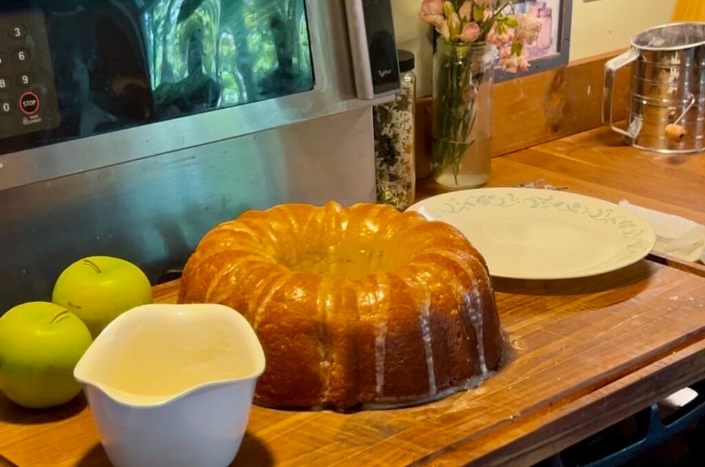 apple Bundt cake on a wooden cutting board, next to extra glaze, green apples, and a plate.