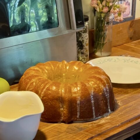 apple Bundt cake on a wooden cutting board, next to extra glaze, green apples, and a plate.