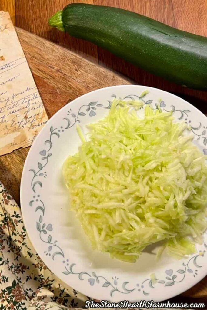Freshly shredded zucchini in a vintage floral bowl alongside a whole garden zucchini and handwritten recipe.