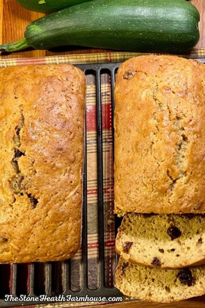 Two golden loaves of homemade zucchini bread with raisins, with fresh green zucchini displayed behind them on a farmhouse kitchen counter.