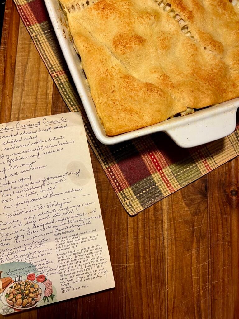 A handwritten chicken casserole recipe next to a freshly baked chicken crescent casserole displayed on a wooden cutting board and plaid country colored table cloth.