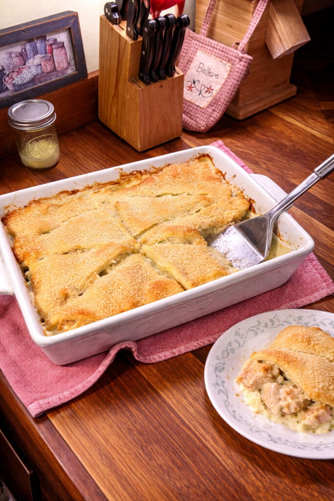 On a wooden counter tip sits the casserole dish with chicken crescent casserole on it. next to it is a piece of the casserole on a plate The casserole is being served. 