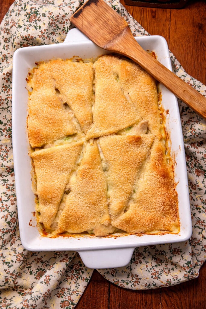On a wooden counter tip sits the casserole dish with chicken crescent casserole on it. next to it is a piece of the casserole on a plate The casserole is being served. 