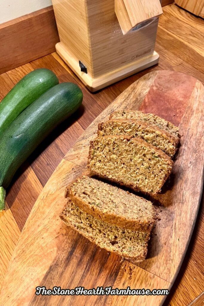 Slices of homemade zucchini bread made with fresh milled flour, served on a wooden cutting board with fresh garden zucchini nearby.