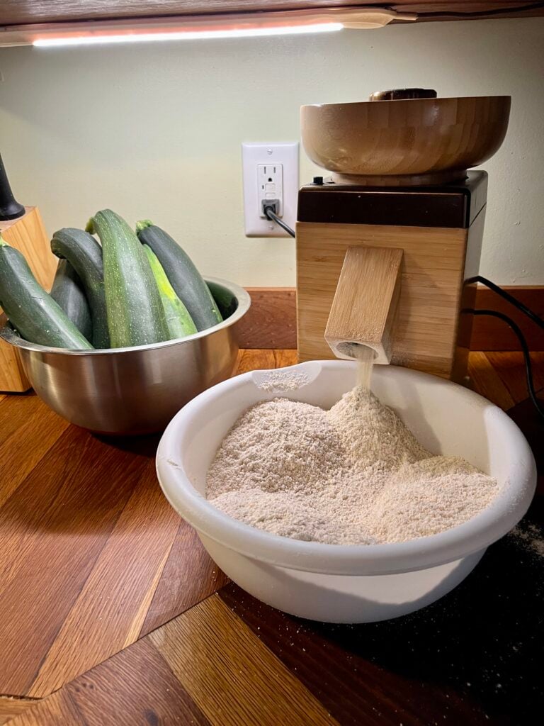 A NutriMill grain mill, milling flour into the bowl to make this healthy fresh milled zucchini bread recipe. 