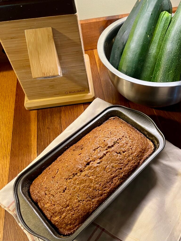 A grainmill, a bowl of ripe zucchinis and a fresh baked loaf of zucchini bread. 