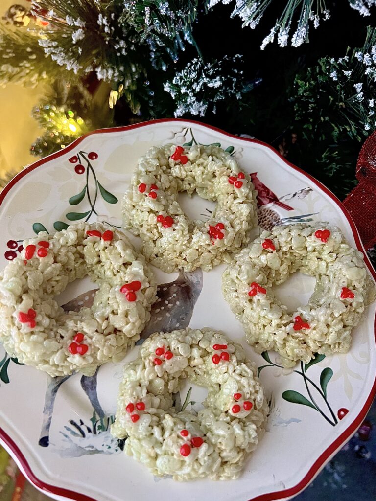Christmas Rice Krispie wreath treats with red holly berry decorations arranged on a holiday plate in front of a decorated Christmas tree.