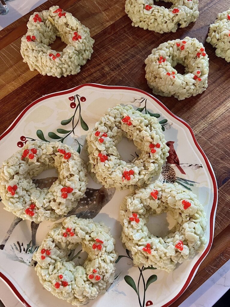 Mini Rice Krispie Christmas wreath treats decorated with holly berries, displayed on a holiday plate and wooden cutting board.