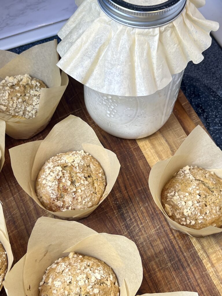 Freshly baked sourdough banana muffins made with sourdough discard, sitting on a wooden cutting board next to a jar of active sourdough starter covered with a parchment lid. Golden muffins with a light crumb topping in parchment liners.
