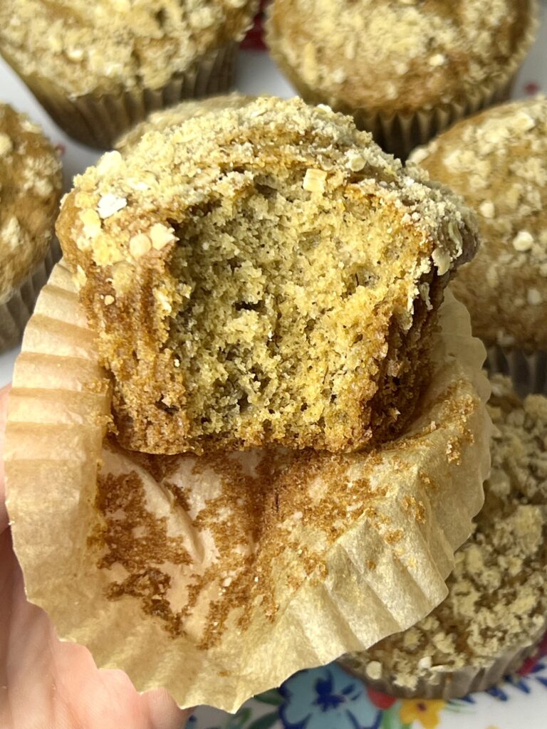 Close-up of a sourdough banana muffin with a crumb topping, showing a soft and fluffy interior after a bite is taken. Homemade sourdough banana muffins with a tender texture and oat crumble sit in the background.
