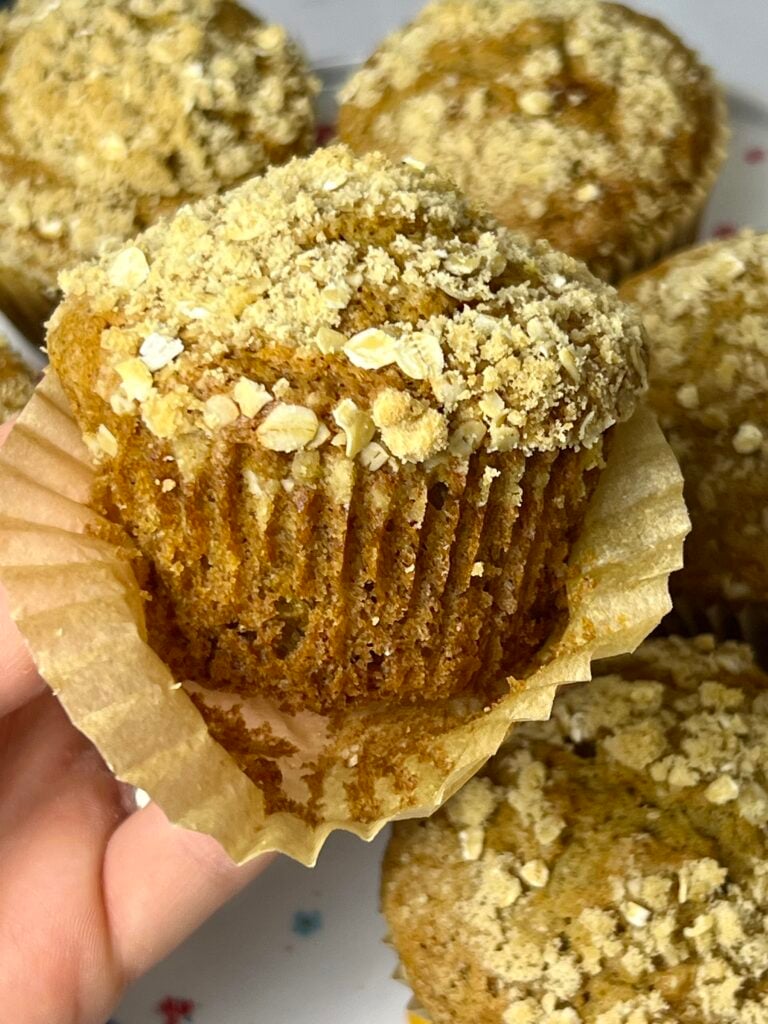 Close-up of a sourdough banana muffin with a crumb topping. Homemade sourdough banana muffins with a tender texture and oat crumble sit in the background.