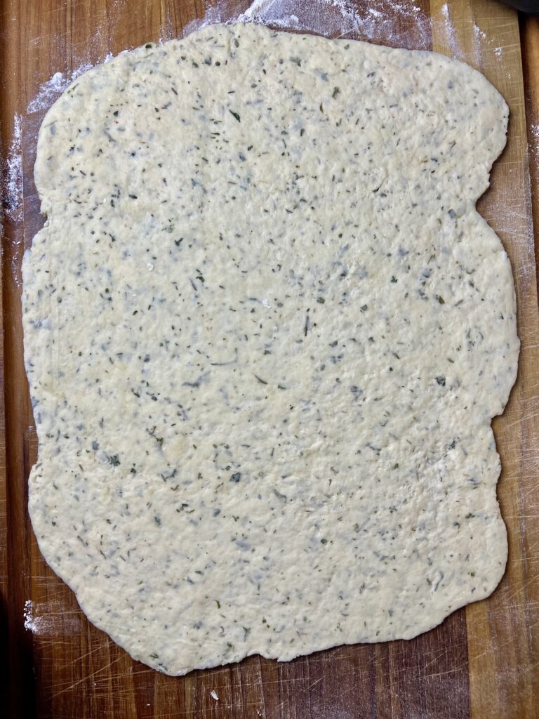 Rolled-out herbed dough on a floured wooden board, ready to be cut and wrapped around sausages for homemade pigs in a blanket.