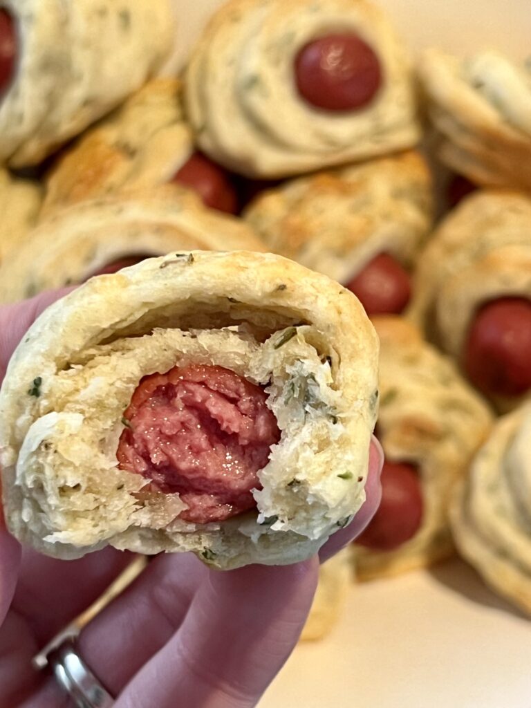 Close-up of a baked pig in a blanket cut open, showing a flaky, herbed homemade dough wrapped around a juicy mini sausage, with more pigs in a blanket in the background.