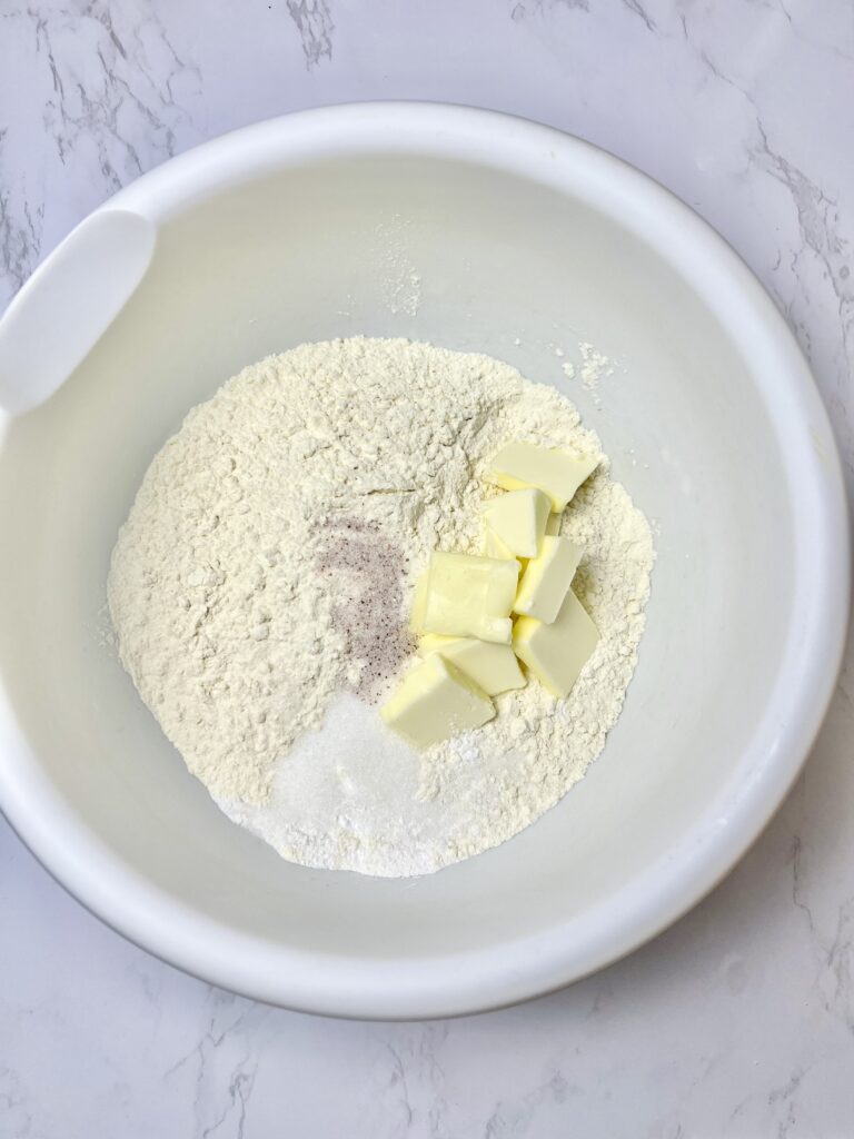 Dry ingredients and cubed butter in a white mixing bowl for pigs in a blanket dough, showing flour, sugar, salt, seasoning, and butter before mixing.