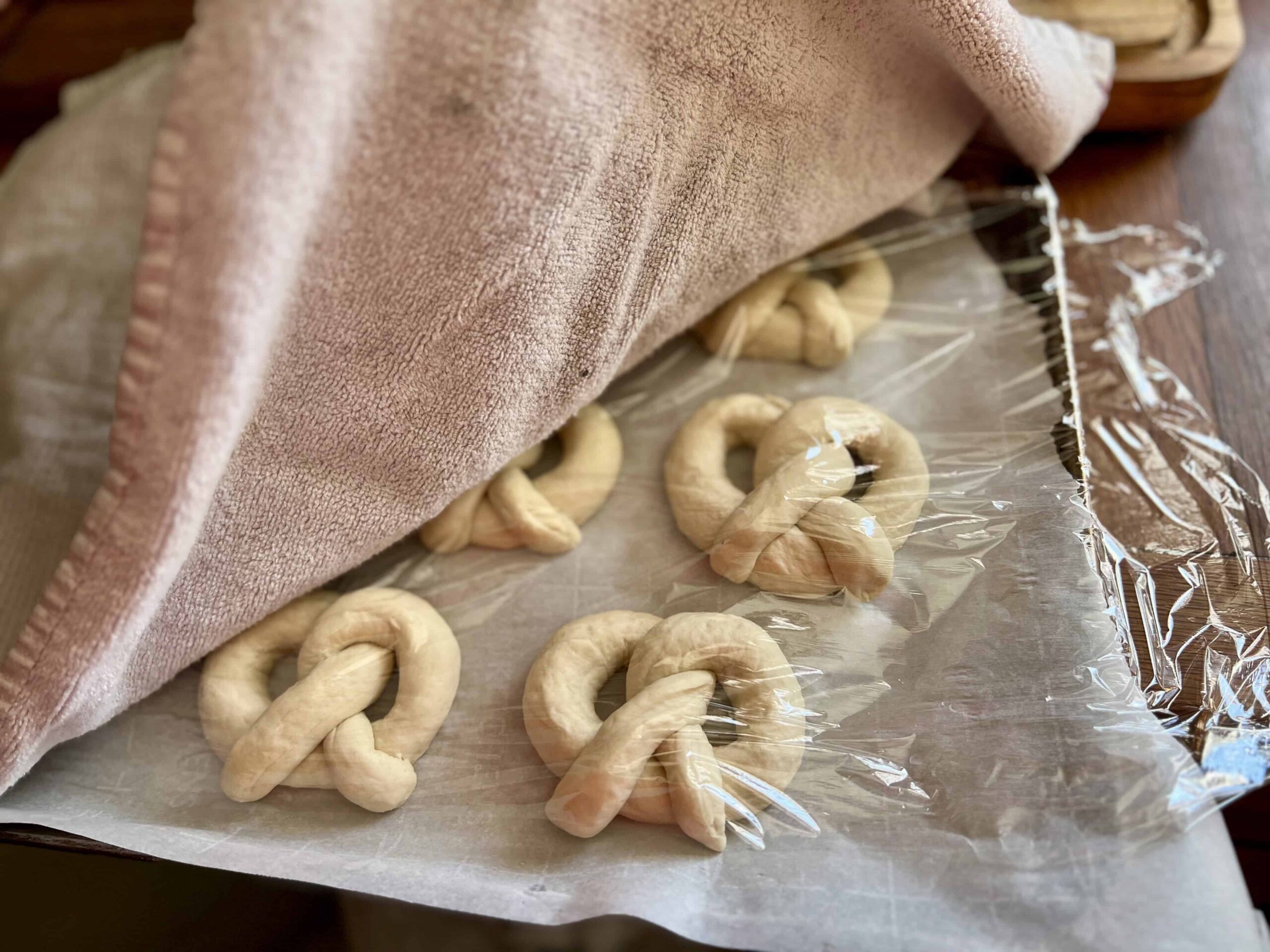 Shaped sourdough soft pretzels resting on a parchment-lined baking sheet, covered with plastic wrap and a towel for rising before baking.