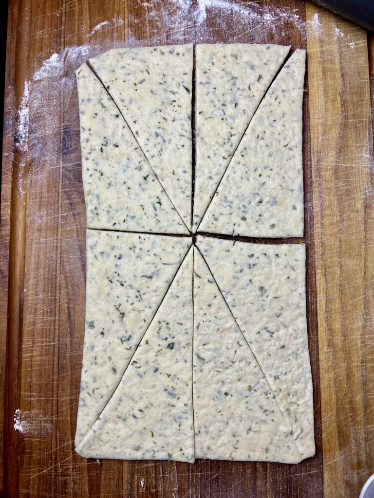 Herbed dough cut into long triangles on a floured wooden cutting board, ready to be wrapped around sausages for homemade pigs in a blanket.